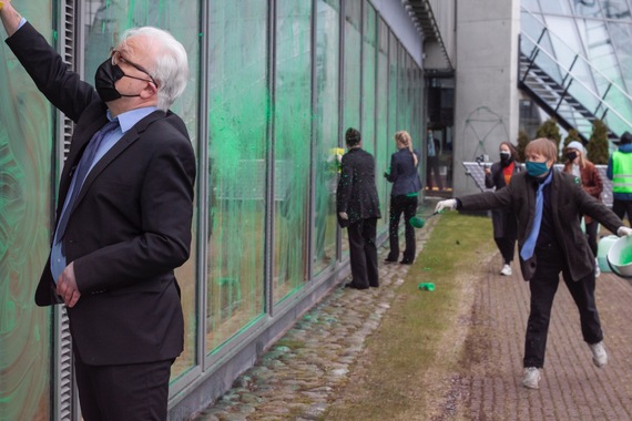 Ulkotila, suuria ikkunoita, joille on levitetty vihreää maalia. Joukko businessasuisia ihmisiä pyyhkii ikkunoita.