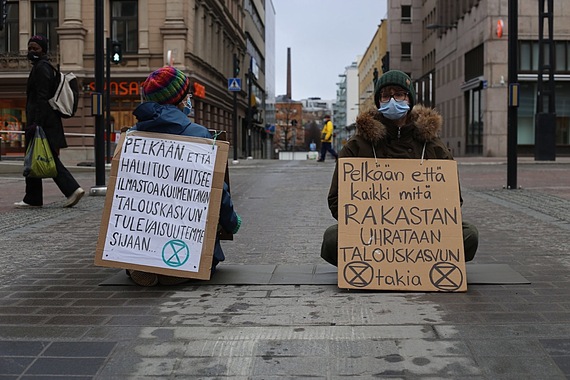 Kaksi ihmistä istuu kaupunkimaisemassa, keskellä autotietä talviasuissa. Oikealla istuva katsoo kameraan, hänellä on kasvoillaan sininen maski, ja edessään kyltti, jossa lukee "Pelkään että kaikki mitä rakastan uhrataan talouskasvun takia". Kyltin alareunasa on kaksi kertaa Elokapinan merkki. Vasemmalla istuva ihminen istuu selin kameraan. Hänellä in kyltti selkäpuolellaan, kyltissä lukee: "Pelkään, että hallitus valitsee ilmastoa kuumentavan 'talouskasvun' tukevaisuutemme sijaan". Kyltin alareunassa on Elokapinan merkki.